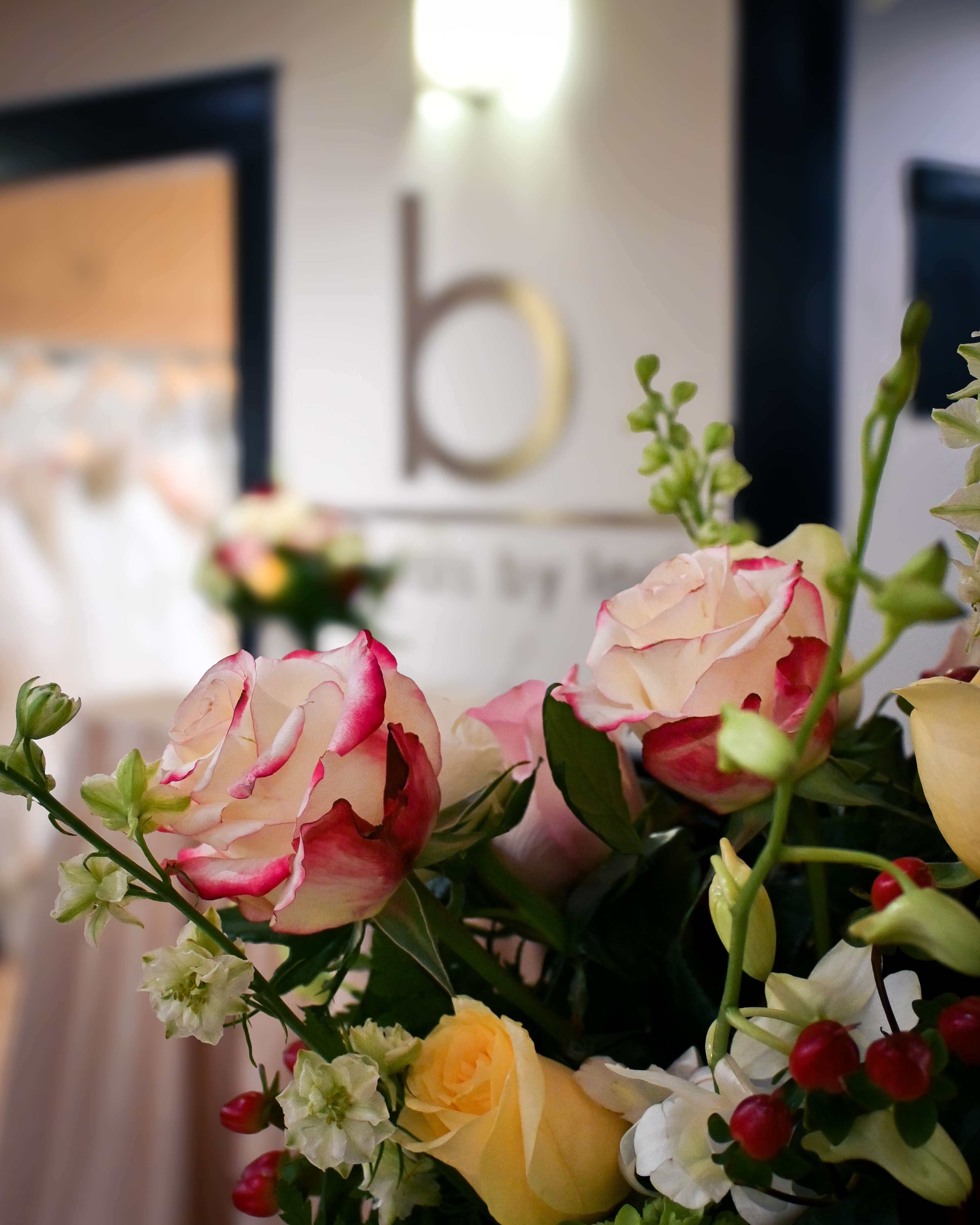 Close-up of a colorful floral arrangement with cream, blush, and yellow roses with pink-tipped petals, small white blossoms, and red berries in the foreground, softly blurred interior of Bridals by Lori in Atlanta visible in the background with the boutique&rsquo;s &ldquo;b&rdquo; logo on the wall.