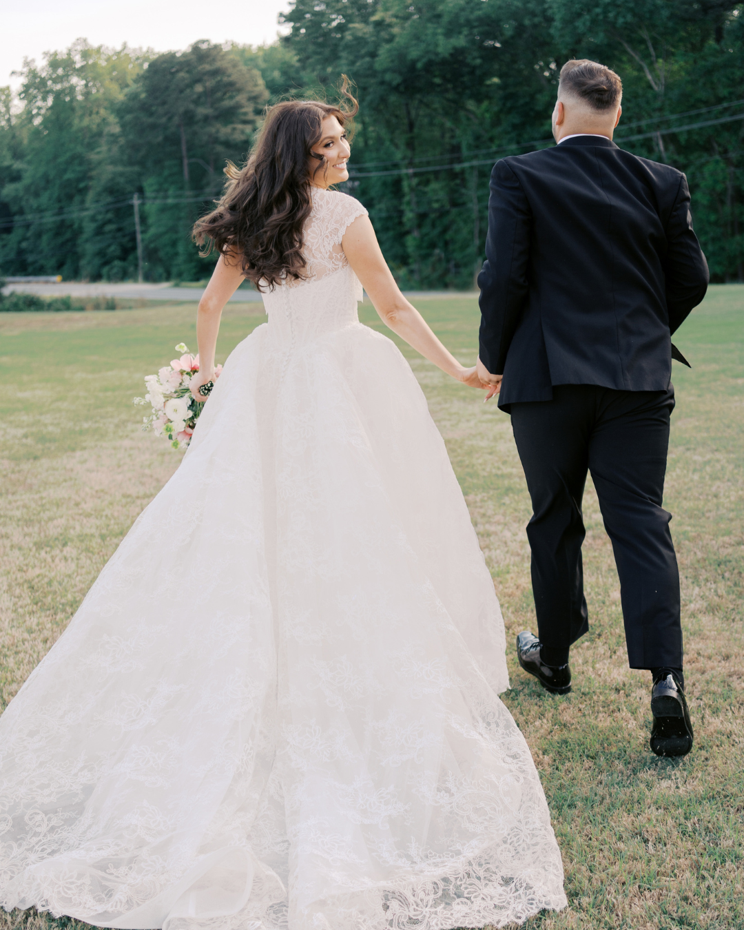 Bride in timeless white lace ballgown wedding dress walking on football field with groom, romantic outdoor portrait showcasing Bridals by Lori designer gown for Southern fall SEC wedding